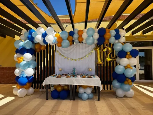 Mesa de doces e arco de balões azuis e dourados ao ar livre.