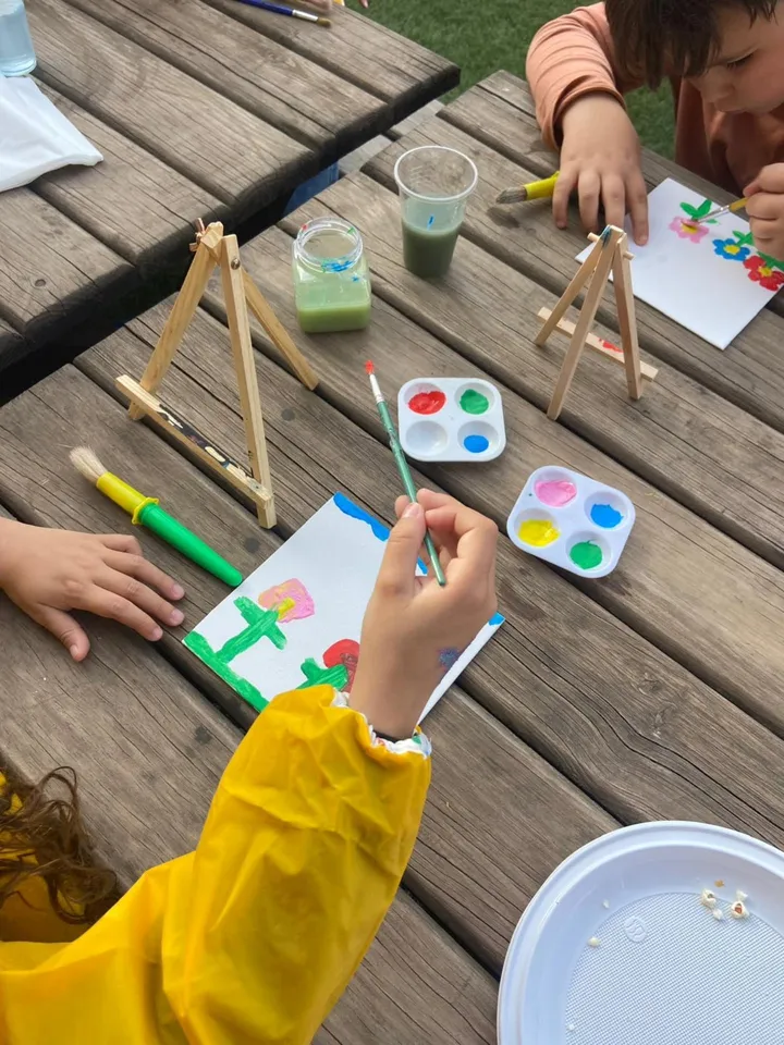 Crianças a pintar telas numa mesa ao ar livre, durante uma atividade da Criar Eventos.